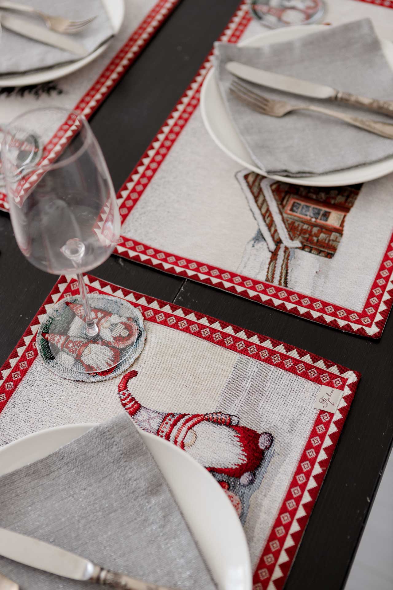 Festive dinner table setup with four Christmas tapestry placemats featuring gnomes and snowy cottage